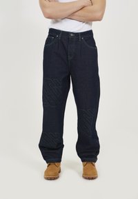 Dark blue denim jeans feature green stitching and a loose fit design. Notable details include large, embossed patterns on the legs.