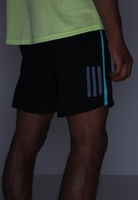 Pantalones cortos deportivos negros con acentos turquesa, que presentan tres rayas verticales grises en la pierna izquierda, fabricados con un material ligero y transpirable.