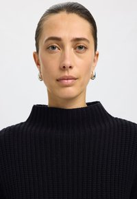 Schwarzer gerippter Pullover mit hohem Kragen, der ein strukturiertes Strickmuster aufweist. Das Model hat kurze Haare und trägt silberne Creolen.