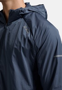 Marine windbreaker jas met capuchon, lichte stof en gestructureerd oppervlak. Reflecterende accenten op de schouders verbeteren de zichtbaarheid.