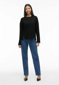 Juoda ilga rankovėmis palaidinė su laisvu kirpimu ir šoninėmis plyšėmis, derinama su aukštu juosmeniu mėlynomis tiesiomis džinsų kelnėmis ir juodais smailiais aukštakulniais.