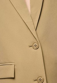 Blazer beige avec une texture lisse, présentant un col à revers, deux boutons et une poche unique sur le côté gauche.