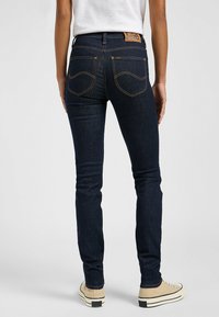 Jeans de denim ajustados en azul oscuro con costuras naranjas en contraste. Presenta dos bolsillos traseros y una etiqueta de cuero en la cinturilla.