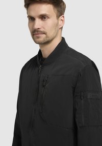 Schwarze Bomberjacke mit einem Reißverschluss vorne, geripptem Kragen und mehreren Taschen, mit einer glatten,matten Textur und minimalistischem Design.