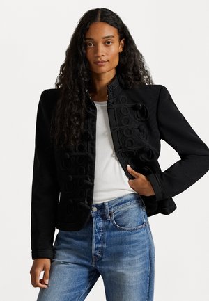 Veste noire avec un design texturé, des accents ronds, un col haut et une silhouette ajustée, associée à un jean bleu clair et une chemise blanche.