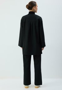 Melna brīvā stila tunika ar augstu apkakli, garām piedurknēm un aizmugurējo krokām; kopā ar melnām plašām biksēm un gaišiem apaviem.