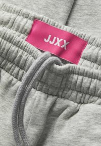 Pantalones de chándal grises con una cintura elástica fruncida y una etiqueta rosa con el texto blanco "JJXX". Textura suave con un diseño casual.