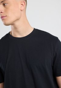 Camiseta de algodón negra con cuello redondo. Mangas cortas y una textura suave, exhibida en un modelo que está ligeramente de espaldas a la cámara.