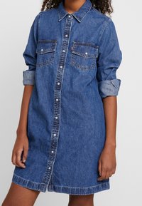 Jeansklänning i blått med knäppning framtill, två bröstfickor och uppvikta ärmar. Har en något figursydd silhuett och fransad nederkant.