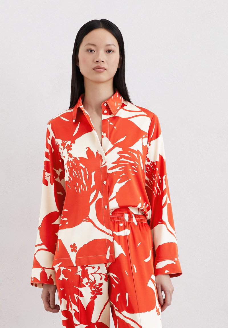 Blouse à manches longues rouge et blanche présentant un motif floral, un col et une fermeture à boutons, fabriquée dans un tissu lisse, associée à un bas assorti.