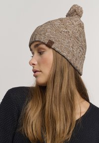 Gehaakte beanie in lichtbruin met een gestructureerd patroon, voorzien van een pompon. Heeft een klein bruin label aan de zijkant.