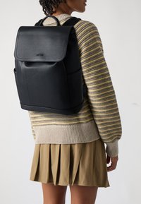 Black leather backpack with a flap closure and two side pockets, featuring a smooth texture and minimalist design.