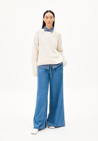 Heller beiger Strickpullover, der über einem Jeanshemd getragen wird, kombiniert mit weit geschnittenen blauen Jeanshosen und weißen Schuhen. Glatte Texturen und lässige Passform.