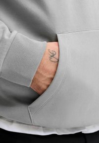 Grauer Pullover mit einem gerippten Bündchen und einer Fronttasche. Die Hand ruht in der Tasche und zeigt ein Tattoo am Handgelenk.