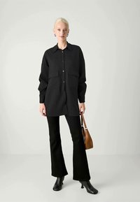 Donna con capelli biondi che indossa una camicia nera a maniche lunghe con bottoni, pantaloni neri svasati, stivali neri alla caviglia e tiene in mano una borsa marrone.
