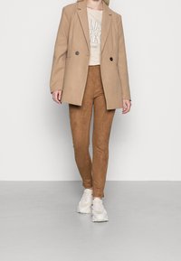 Blazer beige, t-shirt graphique crème, pantalons skinny en daim marron, et baskets de couleur claire. Textures lisses et design ajusté.