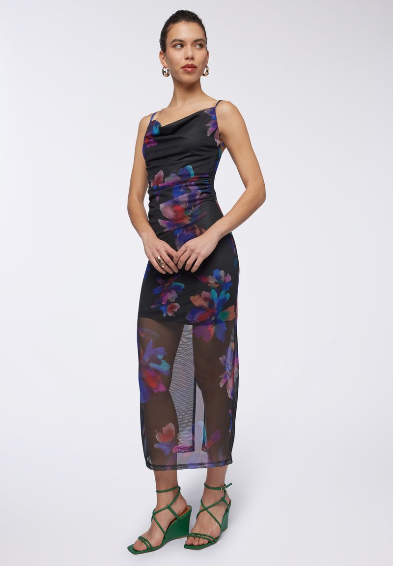 Abito nero con stampa floreale, corpetto arricciato e gonna trasparente. Sandali con tacco a spillo verdi completano il look. Design elegante con colori vivaci.