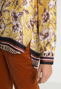 Blouse jaune à motif floral avec une bordure géométrique noire, marron et blanche, associée à un pantalon en velours côtelé couleur rouille et une main détendue sur le côté.