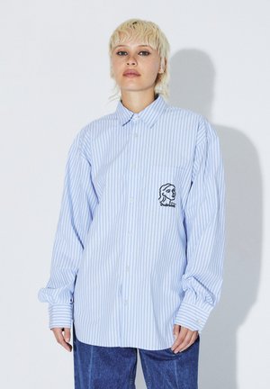 Personne aux cheveux blonds courts porte une chemise boutonnée oversize rayée bleu clair avec un dessin de visage en ligne noire sur la poche poitrine et un jean bleu.