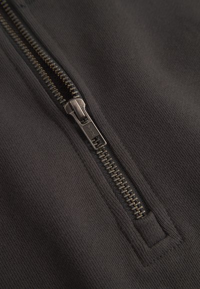 Čierny zipsový odev s hladkou textúrou tkaniny, kovovým zipsom a vystuženým švom okolo zipsu.