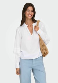 Femme souriante portant un chemisier blanc à manches longues avec des détails en dentelle et un pantalon bleu clair, tenant un sac à bandoulière tissé de couleur sable.