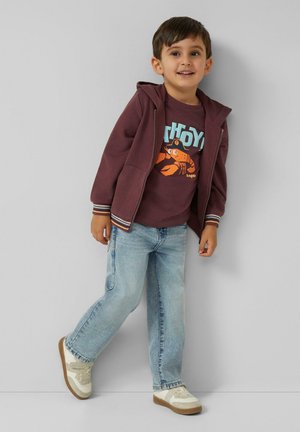 Young boy in maroon hoodie and graphic t-shirt, light blue jeans, and white sneakers, leaning against a gray wall, smiling.
