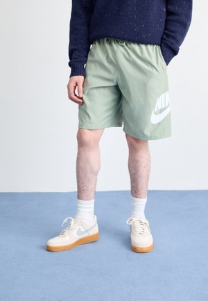 Personne portant un short vert Nike, des baskets blanches avec un swoosh bleu clair, des chaussettes blanches et un pull bleu foncé, debout sur un sol bleu.