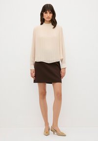 Top plissettato beige con scollatura alta, abbinato a una mini gonna marrone e tacchi beige con un emblema decorativo. Tessuto liscio e leggero.