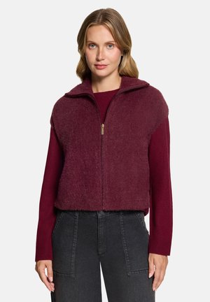 Frau mit blondem Haar trägt eine burgunderrote, flauschige Zip-Weste über einem langärmeligen Pullover und hochgeschnittene schwarze Jeans, steht nach vorne gerichtet.