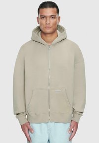 Beige zip-up hettegenser med stor hette, frontlommer, ribbede mansjetter og en myk tekstur. Har en liten logo på lommeområdet.