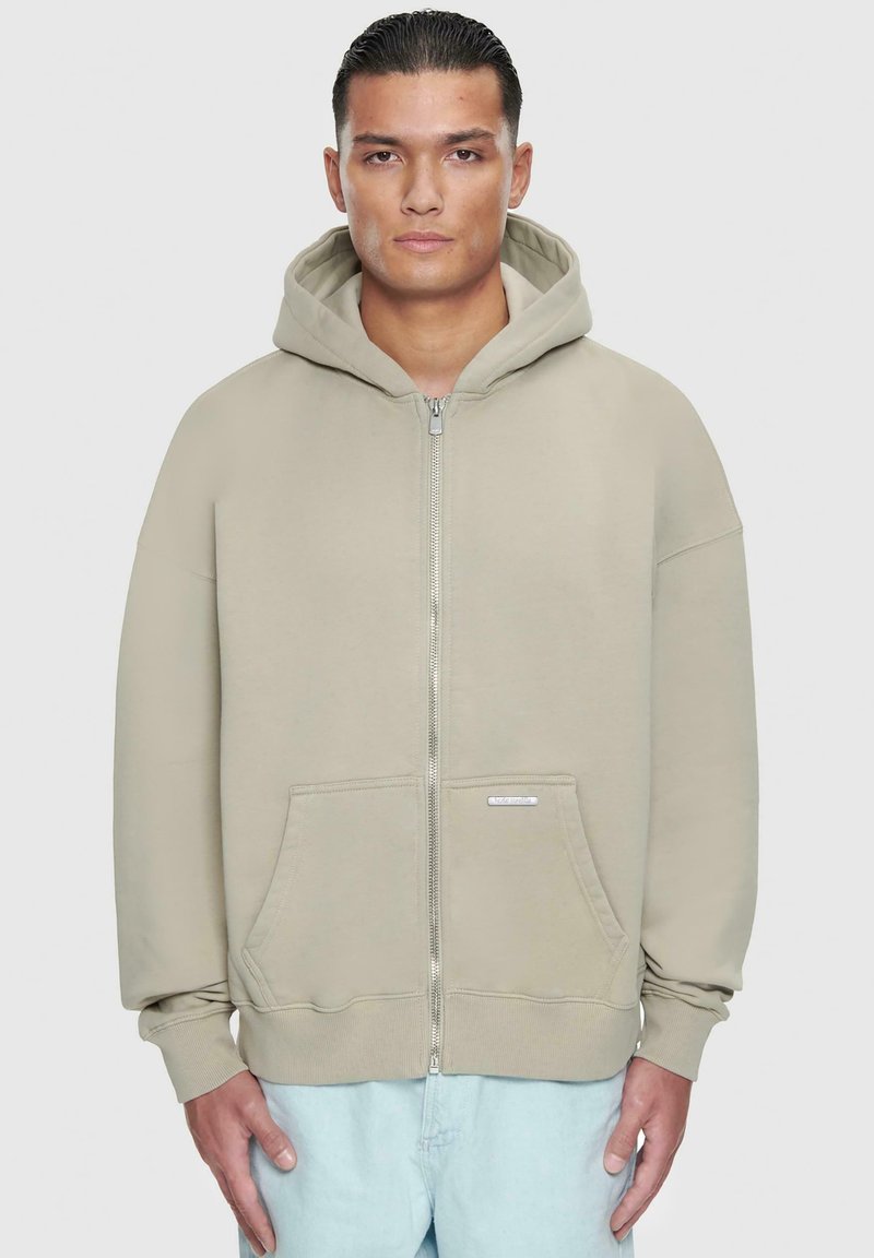 Beige zip-up hettegenser med stor hette, frontlommer, ribbede mansjetter og en myk tekstur. Har en liten logo på lommeområdet.