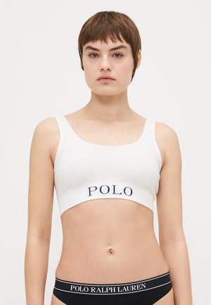 Balts, ribots bralets ar platām tumši zilām siksnām, uz kurām redzams "POLO" logo. Regulējamas siksnas un vienkāršs, minimālisks dizains.