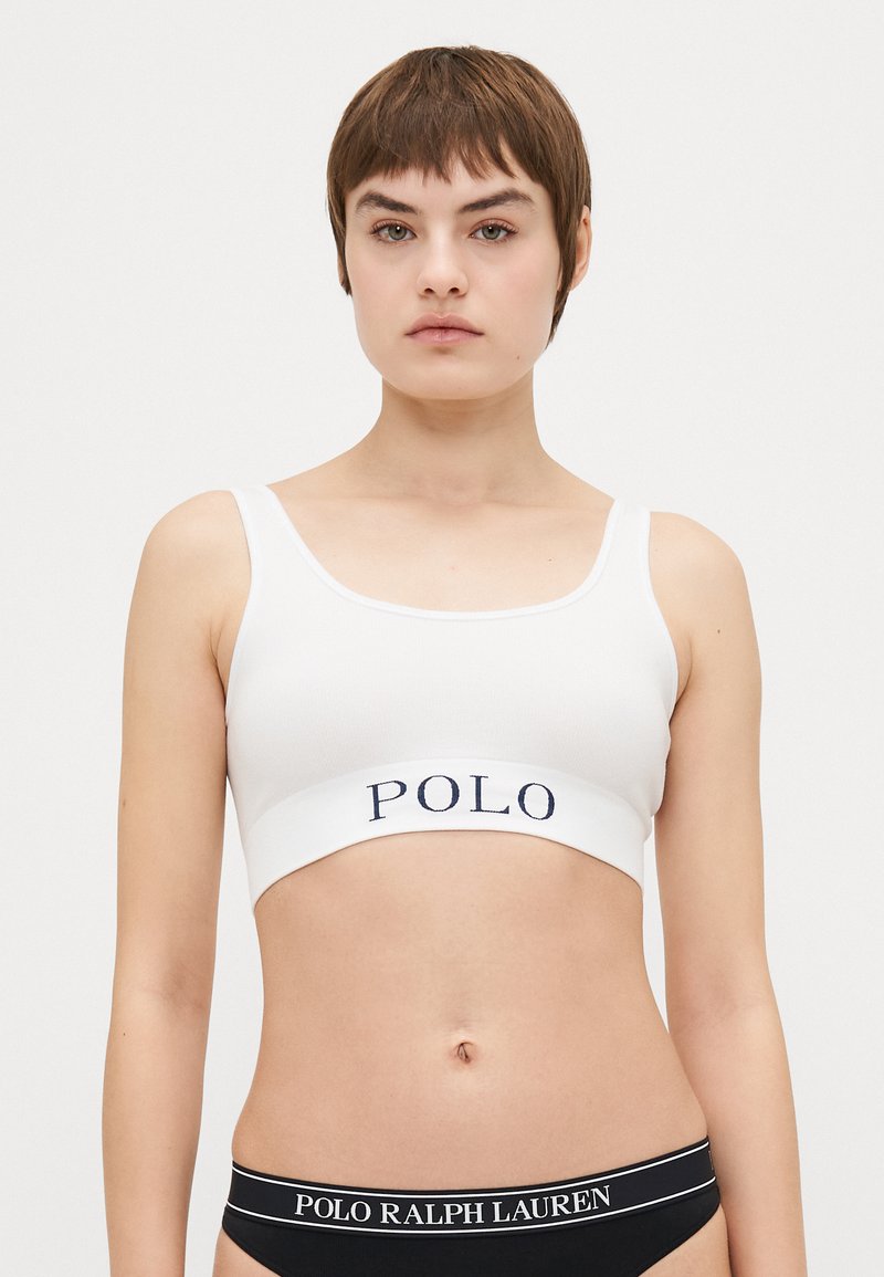 Bela rebrasta braletka z širokim mornarsko modrim pasom, na katerem je izpisan logotip "POLO". Ima nastavljive naramnice in enostaven, minimalističen dizajn.