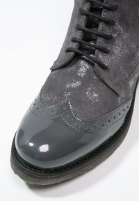Chaussure brogue en suède gris foncé et cuir verni avec perforations détaillées et lacets, montrée en gros plan sur un fond blanc.