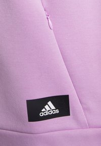 Haut de sport violet clair avec fermeture éclair latérale et écusson logo Adidas noir, en tissu lisse et texturé.