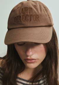Brun kasket med broderet "ART DIRECTOR" i mørkebrun, med en buet skygge og stofstruktur. Justerbar baglukning er ikke synlig.