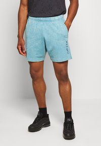Man som bär ljusblå sportshorts med "Just Sport"-text på sidan, svarta sneakers och svarta strumpor, står med ena handen i fickan.