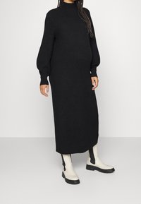 Musta neulottu maksimekko, jossa pitkät, pulleat hihat ja korkea kaulus. Päällä valkoiset nilkkurit, joissa mustat sivupaneelit ja paksu pohja.