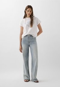 Witte t-shirt met blauwe grafische tekst, gedragen met lichtblauwe wijde jeans. Beide kledingstukken hebben een casual pasvorm en een gladde stoftextuur.