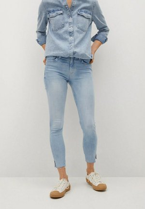 Osoba ubrana w jasnoniebieską koszulę dżinsową włożoną do wyblakłych obcisłych jeansów z postrzępionymi brzegami oraz białe trampki z brązowymi podeszwami.