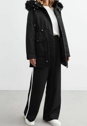 Personne portant un parka noir à capuche en fourrure, chemise blanche, pantalons larges noirs avec bande blanche sur le côté, et baskets blanches sur un fond uni.