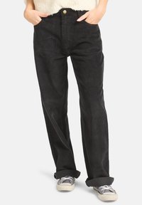 Mains dans les poches, portant un pantalon en velours côtelé noir avec les chevilles retroussées, associé à des baskets grises et blanches usées.