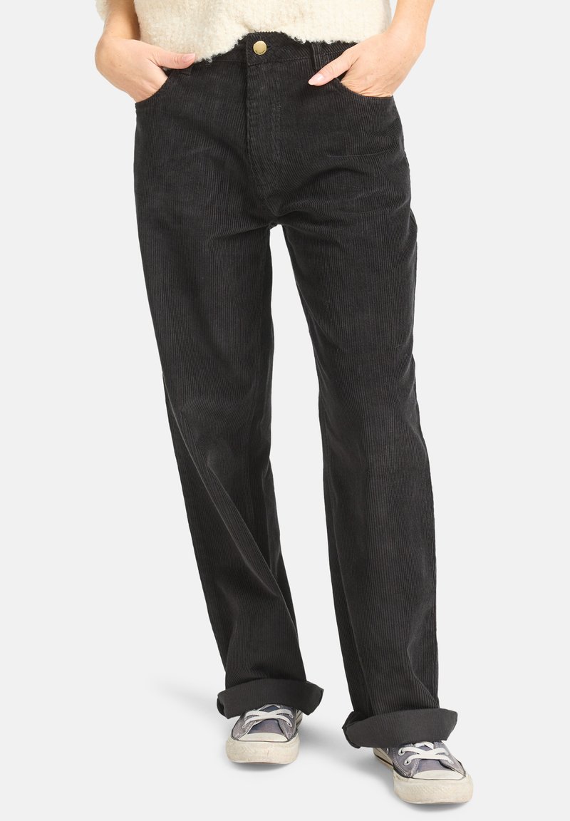 Mains dans les poches, portant un pantalon en velours côtelé noir avec les chevilles retroussées, associé à des baskets grises et blanches usées.
