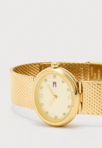 Ρολόι με χρυσή πλέξη λουριού και στρογγυλή κρέμας πρόσοψη. Διαθέτει κρυστάλλινους δείκτες ωρών, λογότυπο του Tom Hilfiger και γυαλιστερό χρυσό περιθώριο.