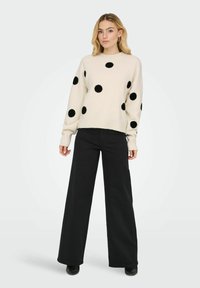 Pull crème à gros pois noirs, col rond et bord-côte. Assorti à un pantalon noir à jambes larges et à des chaussures noires.
