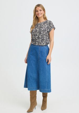 Donna sorridente, indossa una maglietta a maniche corte a fiori bianca e nera, una gonna di denim blu al ginocchio e stivali di camoscio beige al ginocchio su sfondo semplice.