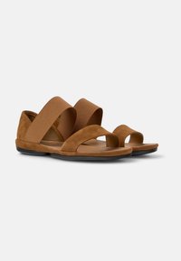 Bruna mockaskinnsandaler med breda elastiska remmar, öppen tå och en platt svart sula. Designen har två korsade remmar för att säkra foten.