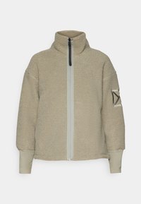 Giacca in pile con zip di colore beige chiaro, con colletto alto e zip grigia a contrasto; presenta una toppa con un design geometrico sulla manica.