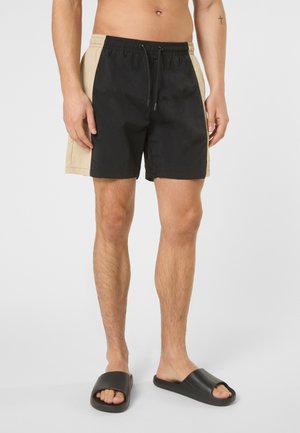 Mand iført sorte og beige farveblok-shorts med elastisk taljebånd og sorte slidesandaler, stående mod en hvid baggrund.