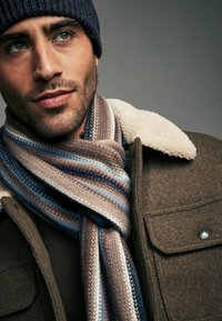 Brown wool coat with shearling collar, featuring a striped knitted scarf in beige, blue, and dark brown. Close-up of face with light stubble.